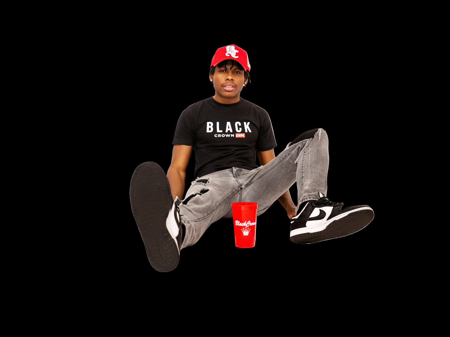 Person wearing a black t-shirt with 'Black Crown' text, gray pants, and black sneakers sitting on a white background with a red cup.