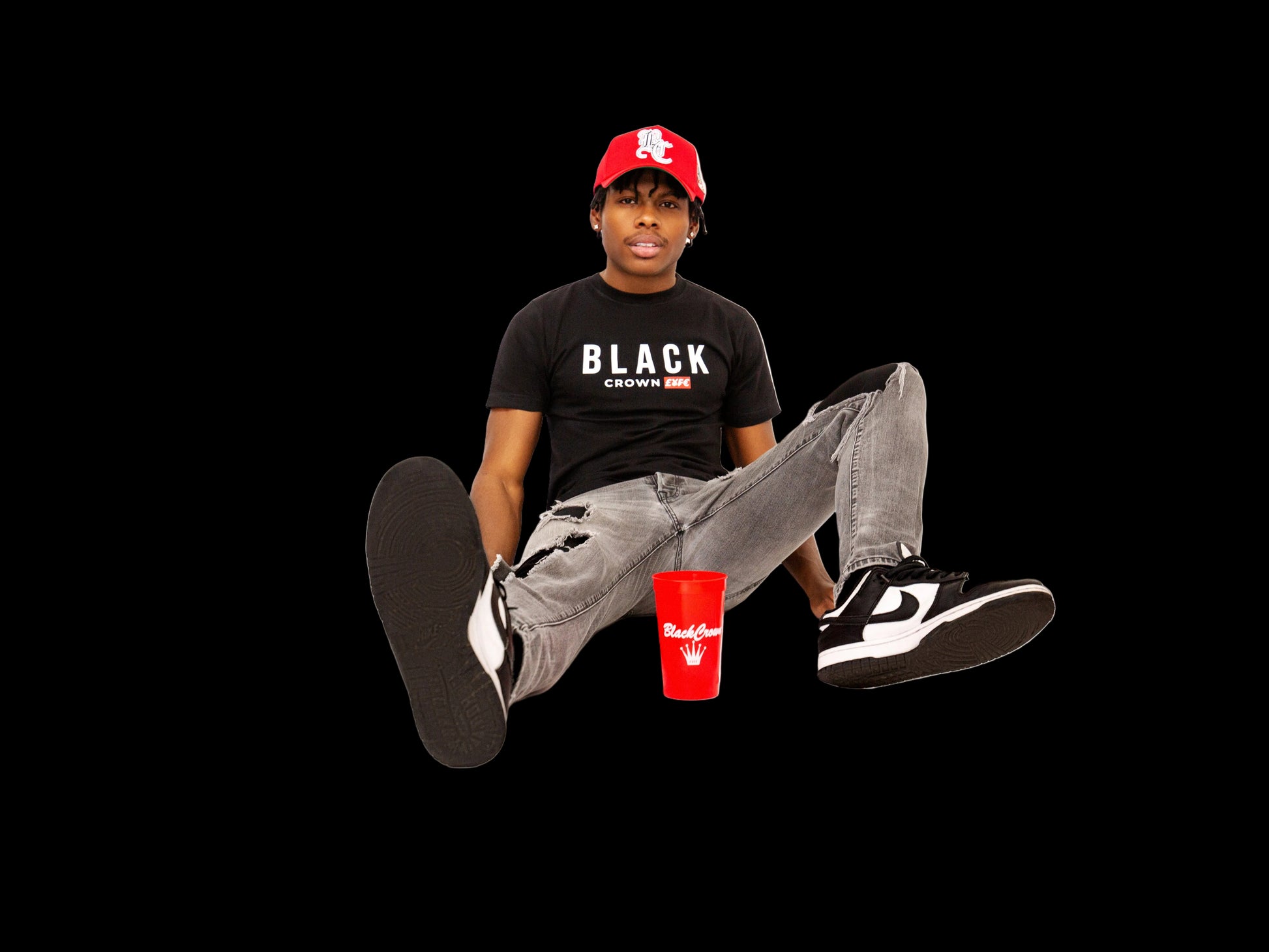 Person wearing a black t-shirt with 'Black Crown' text, gray pants, and black sneakers sitting on a white background with a red cup.